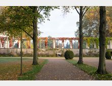 Parkansicht mit einem breiten Weg und Bäumen im Vordergrund, der zu einer langgestreckten Pergola mit überwachsenen roten und grünen Weinranken führt. Im Hintergrund sind eine niedrige Mauer und weitere Bäume zu sehen.