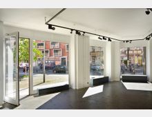 Innenansicht eines leeren, modernen Geschäftsraums mit großen Schaufenstern, die Tageslicht hineinlassen und den Blick auf eine belebte Straßenszene mit parkenden Autos, Fahrrädern und Gebäuden ermöglichen. Die Wände sind weiß, der Boden dunkel, und eine Schienenbeleuchtung ist an der Decke angebracht.