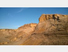 Steilwand einer Sand- oder Erde-Abbaugrube mit sichtbaren geologischen Schichten und spärlichem Pflanzenwuchs an der Oberkante, unter einem klaren, blauen Himmel.