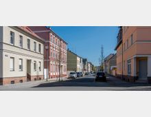 Straßenszene mit zweistöckigen Wohnhäusern, die teils beige, rot und orange gestrichen sind, und geparkten Autos am Straßenrand. Vereinzelte Bäume und Straßenlaternen säumen die gepflasterten Gehwege unter einem klaren blauen Himmel.