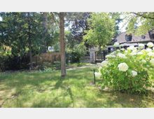 Grünfläche mit Rasen, vereinzelten Bäumen und Sträuchern; im Vordergrund weiße Hortensienbüsche. Im Hintergrund ein eingezäuntes Grundstück mit einem Wohnhaus im Landhausstil, erkennbar an dem dunklen Satteldach und den Dachgauben.