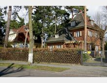 Straßenansicht zweier Einfamilienhäuser in einem Wohngebiet, umgeben von hohen Bäumen. Die Häuser haben Ziegeldächer, rote und gelbe Fassaden, sowie Holzgitterzäune und Einfahrten mit Toren.