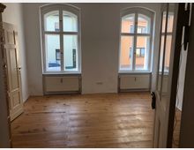 Leerer Raum mit hellen Holzfußböden, hohen weißen Wänden und zwei großen Fenstern mit Rundbögen, die viel Tageslicht hereinlassen. Unter den Fenstern befinden sich Heizkörper, und links ist eine geöffnete Innentür sichtbar.