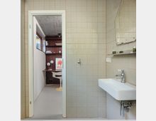 Small tiled bathroom with light square wall tiles, a wall-mounted white sink and chrome faucet beneath a shelf and mirror. An open doorway leads to an adjacent room with built-in dark wood shelving, a desk area, and a window.