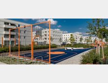 Moderner Wohnbereich mit einer Basketballanlage, die von einem orangen Zaun umgeben ist. Im Hintergrund sind weiße Mehrfamilienhäuser, Grünflächen sowie ein Spielplatz mit Klettergerüst zu sehen.