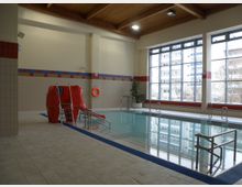 Innenraum eines Hallenbads mit einem rechteckigen Becken, umgeben von Fliesenboden. Am Beckenrand stehen Leitern, eine rote Kinderrutsche und ein Rettungsring, während große Fenster auf der rechten Seite den Blick auf Gebäude und Bäume freigeben.