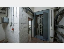 Ein Technikraum mit weiß gestrichenen Ziegelwänden, einem offenen Schaltschrank mit sichtbaren Kabeln und Schaltanlagen sowie Kabeln, die entlang der Wände und Kabeltrassen verlaufen. Eine offene Tür führt in einen angrenzenden Raum mit weiterem technischen Equipment.