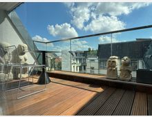 Eine moderne Terrasse mit Holzboden, Glasgeländer und Blick auf umliegende Gebäude mit Schrägdächern. Auf der Terrasse stehen Drahtstühle, ein kleiner Tisch und dekorative Steinskulpturen.