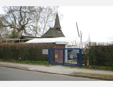 Gebäudeeingang mit blauer Gittertür, hinter der eine Hecke und ein Gebäude mit spitzem Holzdach sichtbar sind. Im Hintergrund stehen kahle Bäume, und der Himmel ist bewölkt.