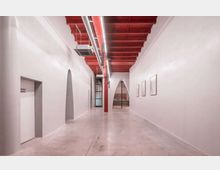 Ein moderner Innenraum mit weiß gestrichenen Wänden, einem roten Deckenträger aus Holz und sichtbaren Lüftungsrohren. Eine Glaswand mit schwarzen Rahmen ist im Hintergrund zu sehen, daneben befinden sich bogenförmige Durchgänge und minimalistische Wanddekorationen.