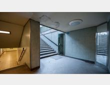 Unterführung mit nackten Wänden aus kleinen Fliesen, abgeblättertem Putz und Betonboden. Zwei Treppen führen nach oben, eine mit Metallgeländer, während auf der linken Seite ein Flur mit Fliesenboden zu sehen ist.