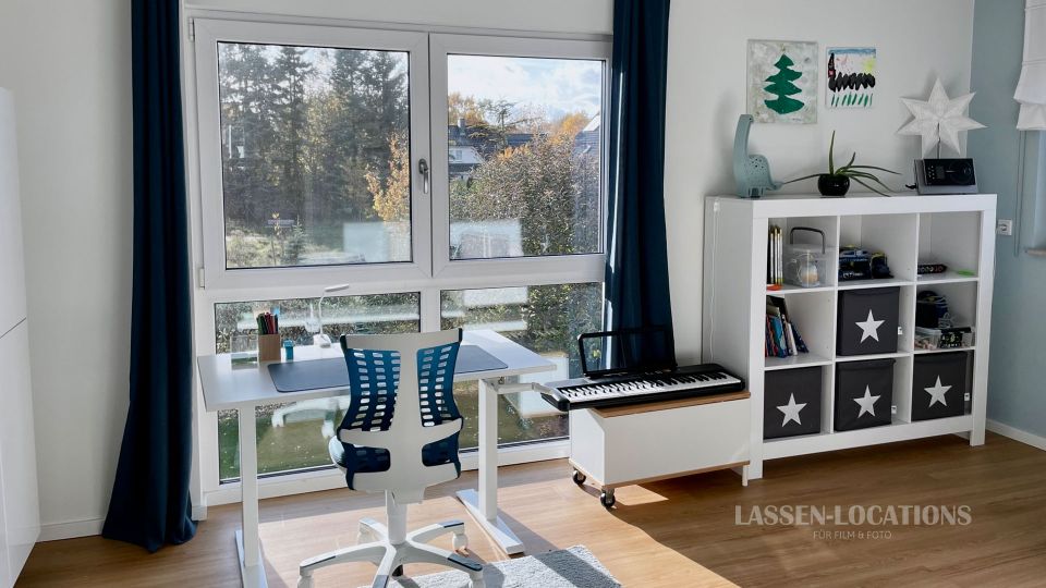 Helles Arbeitszimmer mit großem Fenster und dunklen Vorhängen, davor ein weißer Schreibtisch mit Bürostuhl und Schreibutensilien. Rechts stehen ein weißes Regal mit schwarzen Stoffboxen, ein kleines Keyboard auf einem niedrigen Rollschrank sowie Wanddeko und eine Pflanze.