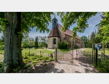 Historische Kirche mit einem rechteckigen Kirchenschiff aus Naturstein und einem Turm mit spitzem Dach, umgeben von Bäumen und einem kleinen Garten. Im Vordergrund befindet sich ein schmiedeeisernes Tor und ein gepflasterter Weg, der zur Kirche führt.