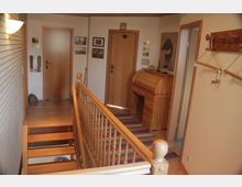 Flur in einem Wohnhaus mit Holzboden, einer Holztreppe mit Geländer und mehreren Türen an den Wänden. An der Wand hängen Bilder und Dekorationen, ein Schreibsekretär steht präsentiert, und ein bunter Läufer bedeckt den Boden.