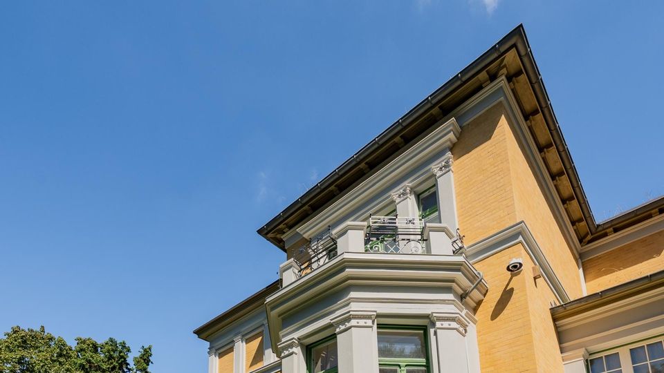 Außenansicht eines mehrstöckigen historischen Wohnhauses mit gelblicher Fassade, weißen Gesimsen und Erkern sowie einem kleinen Balkon mit Metallgeländer. Im Hintergrund sind blauer Himmel und Baumkronen zu sehen.
