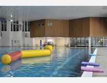 Innenansicht eines Hallenbads mit einem großen Becken, hellblauem Wasser und einer gelb-roten Schwimmrolle auf der Wasseroberfläche. Der Raum hat eine Holzverkleidung an den Wänden, hohe Fenster, eine helle Decke und Sitzbänke am Beckenrand.