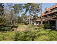Großer Garten mit Rasenfläche, Bäumen und einem Teich, umgeben von dichter Vegetation. Rechts im Bild steht ein großes Wohnhaus mit rotem Ziegeldach und mehreren Fenstern; die Umgebung wirkt ländlich und ruhig.