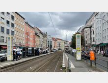 Straßenbahnhaltestelle in einer urbanen Umgebung mit mehrstöckigen Wohn- und Geschäftshäusern in verschiedenen Farben entlang der Straße. Die Haltestelle verfügt über Wartehäuschen und Informationssäulen, während Straßenbahnschienen durch die Mitte der Straße verlaufen.
