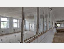 Ein leerstehender, gefliester Innenraum mit weißen Wänden und großen, rechteckigen Fenstern. Im Vordergrund sind Trennwände mit Glasfenstern und beschädigten Rahmen, der Boden ist mit quadratischen Fliesen ausgelegt.