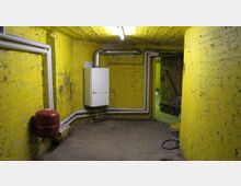 Ein Kellerraum mit gelb gestrichenen Backsteinwänden, sichtbaren Rohren und einem weißen Heizsystem an der Wand. Am Boden stehen ein rotes Ausdehnungsgefäß sowie ein gelber Farbeimer, und im Hintergrund ist eine Holztür zu sehen.