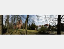 Panorama einer parkähnlichen Umgebung mit Bäumen, einer Wiese und einem roten Backsteingebäude im neugotischen Stil, das eine Kirche darstellt. Im Hintergrund sind weitere Wohnhäuser mit roten Dächern zu sehen, die von Bäumen und Sträuchern umgeben sind.
