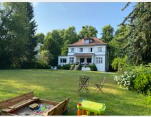 Großer Garten mit Rasenfläche und Spielplatzbereich, im Hintergrund ein weißes zweistöckiges Haus mit rotem Ziegeldach und Balkon. Umgeben von hohen Bäumen und Sträuchern, sind auch Gartenmöbel und ein Sandkasten im Vordergrund sichtbar.