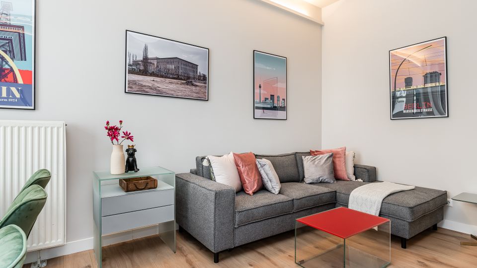 Modernes Wohnzimmer mit grauem Ecksofa und farbigen Kissen, kleinem Glastisch mit roter Ablage sowie hellem Holzboden. An den weißen Wänden hängen mehrere gerahmte Poster; daneben stehen ein schmaler Heizkörper und ein kleiner Beistelltisch mit Vase.