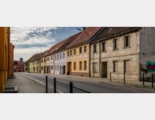 Straßenszene mit Reihenhäusern in einer ruhigen Stadt. Die Gebäude haben bunte Fassaden, während eines der Häuser links ungepflegt wirkt; Bürgersteig mit Kopfsteinpflaster und ein Zaun säumen die Straße.