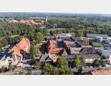 Luftaufnahme eines weitläufigen Industrie- und Fabrikgeländes mit mehreren Backsteinhallen und roten Ziegeldächern, umgeben von vielen Bäumen. Rechts sind technische Anlagen mit Rohrleitungen und runden Tanks zu sehen, im Hintergrund Waldflächen und eine Siedlung mit einem markanten Turm.