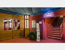 Foyer eines Veranstaltungsraums mit roten Wänden und Teppichboden, einer hölzernen Doppeltür mit Glaseinsätzen, einer Treppe mit rotem Teppich und Geländer sowie einem Prospektständer und einem großen Lautsprecher. Die Decke ist mit blauen Stoffbahnen bespannt.