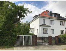 Zweistöckiges weißes Wohnhaus mit rotem Schrägdach und mehreren Fenstern, umgeben von Bäumen und Hecken. Vor dem Haus befindet sich ein gepflasterter Gehweg sowie ein Zaun mit zwei Toren aus Metall, teilweise von Backsteinsäulen gestützt.