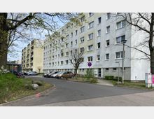 Wohngebäude mit vier Etagen in einer Wohnsiedlung, erkennbar an weißen und beige-grauen Fassaden mit rechteckigen Fenstern. Vor dem Gebäude befinden sich ein kleiner Parkplatz mit geparkten Autos, eine Straße, Gehwege, sowie Bäume und Sträucher.