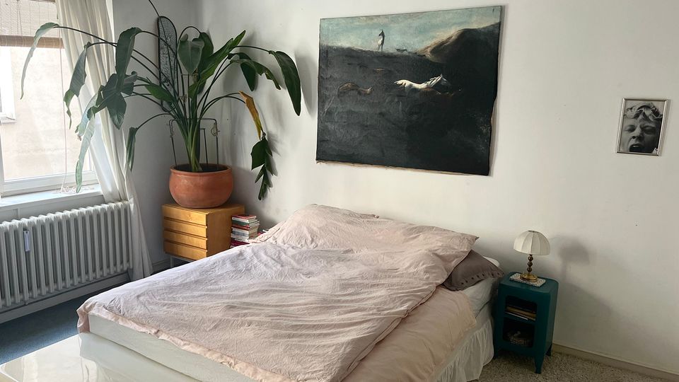 Bright, minimalist bedroom with a low bed and pale bedding, a teal nightstand with a small lamp, and white walls decorated with framed artwork. A large potted plant sits on a wooden dresser beside a window with sheer curtains and a radiator below.