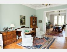 Wohnzimmer mit pastellgrünen Wänden, verziertem Stuck an der Decke und einer Einrichtung im klassischen Stil mit antiken Holzmöbeln, gestreiften Polstermöbeln und Teppichen mit orientalischen Mustern. Im Hintergrund ist ein angrenzender Essbereich mit großem Esstisch und mehreren Stühlen zu sehen, der von Tageslicht durch große Fenster erhellt wird.