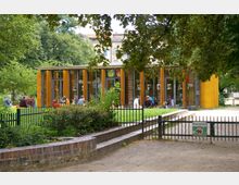 Ein moderner, eingeschossiger Pavillon mit großen Glasfenstern und gelben Holzelementen, umgeben von Grünflächen und Bäumen. Im Vordergrund befindet sich ein Metallzaun mit einem Schild „Hunde verboten“, und Menschen sitzen auf der Wiese vor dem Gebäude.