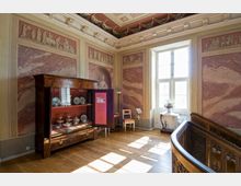 Ein Innenraum eines historischen Museums mit aufwendig verzierten Wänden, die einen Marmoreffekt und klassische Reliefs zeigen. Zu sehen sind ein Holzvitrinenschrank mit ausgestelltem Porzellan, ein Fenster mit hellem Tageslicht sowie Möbel wie ein kleiner Tisch mit Blumenschmuck.