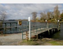 Eine kleine Holzsteganlage mit Metallgeländern ragt in einen See, umgeben von kahlen Bäumen und Häusern am Ufer. Im Vordergrund führt ein gepflasterter Weg zur Anlegestelle, und ein Rettungsring ist an einer Metallhalterung befestigt.