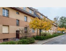 Straßenszene mit einer Reihe von mehrstöckigen Wohngebäuden in beigefarbenem Putz und rotbraunem Ziegeldach. Vor den Gebäuden stehen Bäume mit herbstlich gelbem Laub, umgeben von gepflegten Hecken und Gehwegen.