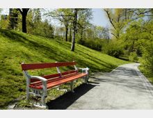 Ein Park mit einer geschwungenen, asphaltierten Gehwegfläche, die von grünen Wiesen und Bäumen gesäumt ist. Im Vordergrund steht eine rot-weiße Parkbank am Wegesrand.