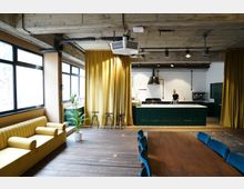 Großzügiger, modern gestalteter Raum mit industriellen Elementen, bestehend aus einer offenen Küche mit grünen Schränken, einem großen Holztisch und einer Sitzgruppe mit gelbem Sofa. Der Raum hat Sichtbetondecken, große Fenster, goldene Vorhänge und Holzboden, ergänzt durch dekorative Pflanzen.