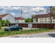 Wohngebiet mit einem Parkplatz im Vordergrund, auf dem ein dunkles Auto steht. Dahinter eine Reihe alter Garagen mit braunen Türen, Wohnhäuser mit roten Ziegeldächern und ein Funkmast im Hintergrund.