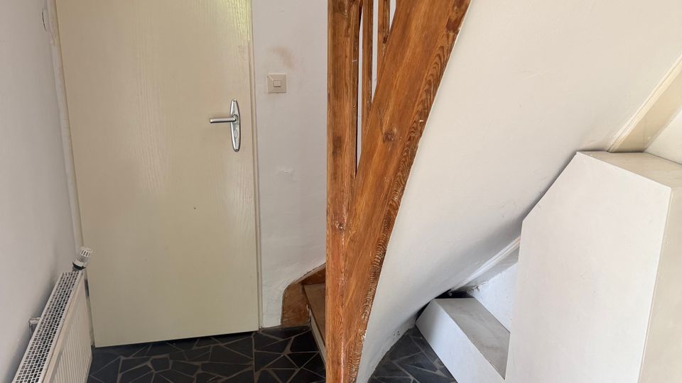 Kleiner Innenflur mit einer geschlossenen, hellen Tür und einer Holztreppe mit Geländer; links steht ein Heizkörper an der Wand. Der Boden ist mit unregelmäßigem, dunklem Steinmosaik gefliest.
