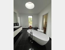 Modernes Badezimmer mit freistehender Badewanne in der Mitte, schwarzen glänzenden Fliesen und einer großen Fensterfront mit Blick ins Grüne. Links befinden sich ein Waschbecken mit schwarzer Steinplatte und weiße Schränke, oben ein großes rundes Oberlicht.