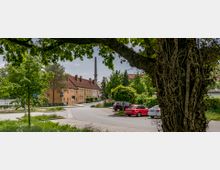 Straßenszene in einem Wohngebiet mit mehrstöckigen, gelblichen Reihenhäusern mit roten Ziegeldächern, umgeben von Bäumen und grünem Bewuchs. Im Hintergrund ragt ein hoher Schornstein auf, während im Vordergrund ein großer Baum sowie geparkte Autos entlang der Straße sichtbar sind.