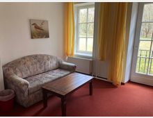 Wohnzimmer mit einem gepolsterten, gemusterten Sofa, einem Holztisch und rotem Teppichboden. Große Fenster mit gelben Vorhängen bieten Blick auf einen Garten, und eine Tür führt ins Freie.