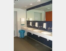 Öffentliche Waschräume mit einer langen weißen Waschbeckenreihe unter einem großen Spiegel, blau gefliesten Wänden und einem grauen Fliesenboden. Ein Mülleimer mit einem blauen Plastiksack steht links neben der Waschbeckenreihe.