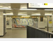 Unterführung in einer U-Bahn-Station mit gefliesten Wänden, einem geschlossenen und mit Graffiti besprühten Rolltor sowie Leuchtstofflampen an der Decke. Ein Wegweiser weist den Ausgang zur Bundesallee und Landhausstraße aus.