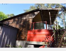 Holzhaus mit vertikaler Holzverschalung, einer roten Veranda mit Glasverzierung und einem schrägen Wellblechdach. Die Umgebung ist von Bäumen und Pflanzen geprägt, unter einem klaren blauen Himmel.