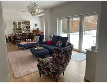 Offener Wohn- und Essbereich mit blauem Ecksofa, Hocker und gemustertem Sessel auf Teppich sowie einem langen Esstisch mit Stühlen und Vitrinenschrank im Hintergrund. Große bodentiefe Glas-Schiebetüren führen in einen verschneiten Garten mit Bäumen.