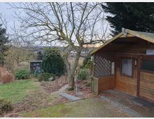 Garten mit einer kleinen Holzlaube rechts und einem weiteren kleinen Schuppen im Hintergrund. Die Umgebung ist von Bäumen, Sträuchern und Wiese geprägt, mit teilweise kahlen Ästen und einer winterlichen Atmosphäre.